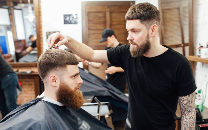 Man met een op Caesar geïnspireerde drop fade en een grote bossige baard kijkt vooruit terwijl hij zijn haar laat knippen.