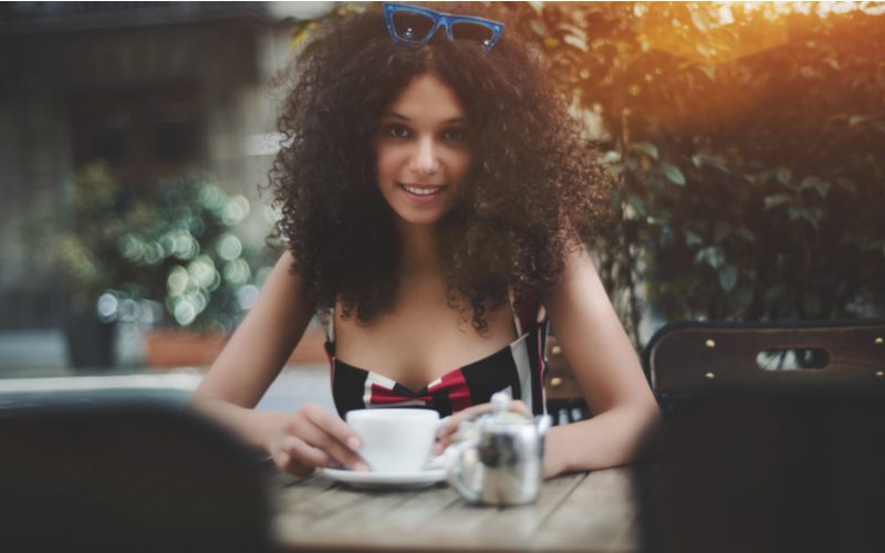 Mooie lichtgekleurde vrouw in een jurk met patroon, coily haar en een bril op haar hoofd houdt een espresso glas vast.