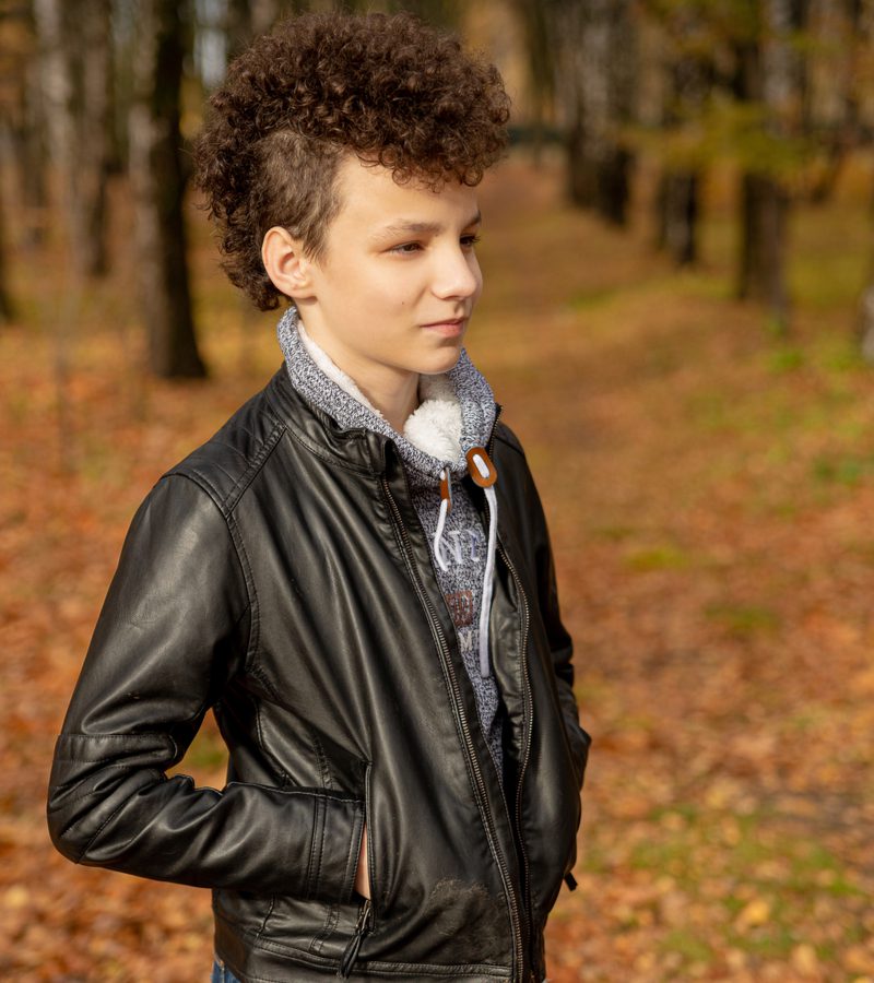 Man in een leren jas die een Curly Mohawk Mullet Fade draagt terwijl hij in een bos staat in de herfst.