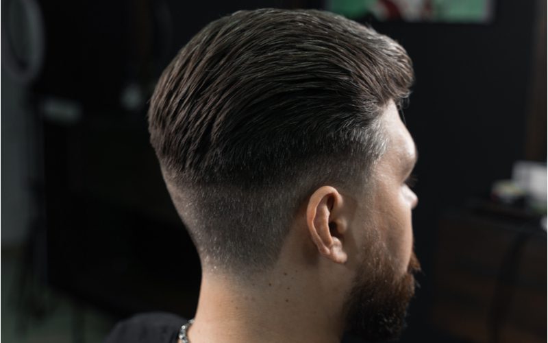 Zij- en achterprofiel van een man met een slicked-back low fade kapsel in een verduisterde kamer.