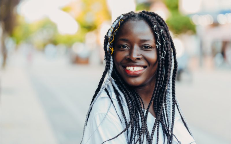 Vrouw met box braids staat in een wit hemd buiten.