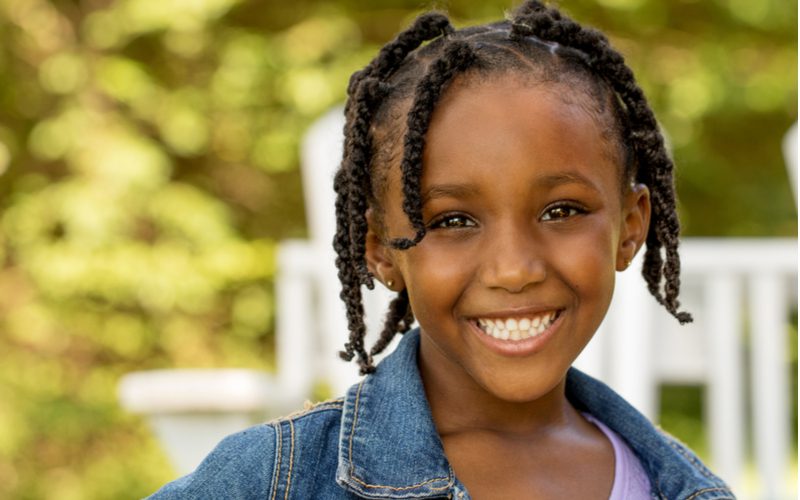 Kindvriendelijke box braids op een meisje met kralen in haar haar dat voor een wit hek staat.