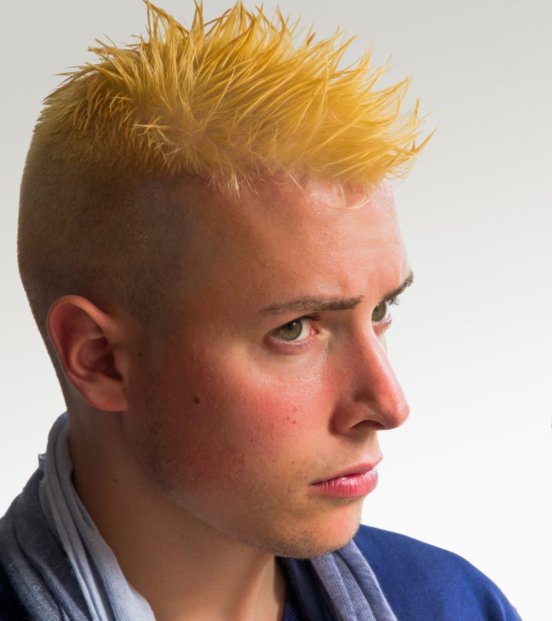 Bleach-Blonde Faux Hawk op een kerel met oranje haar die een blauw hemd draagt met een sjaal en naar rechts van de lezer kijkt.