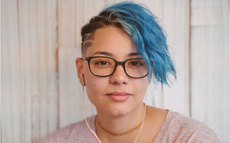 Liberale lesbische vrouw met blauw haar en een bril met een geschoren kant die het punkkapsel rockt en naar muziek luistert op een gouden hoofdtelefoon.