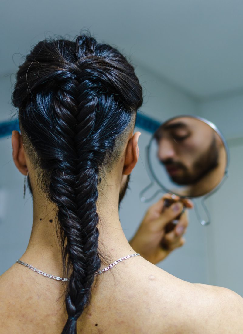 Jonge man met een vissenstaart vlecht voor mannen kijkt naar zijn smerige baard in de spiegel