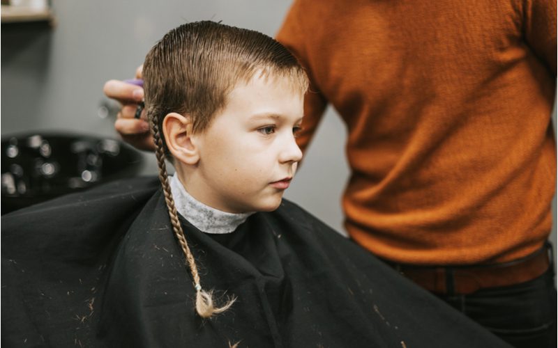 Jongen met vlechten voor mannen zittend in een kappersstoel naast een jongen in een oranje trui