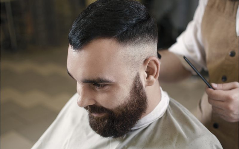 Man draagt een nette undercut fade terwijl hij glimlachend in een kappersstoel zit en een zeer dikke baard draagt.