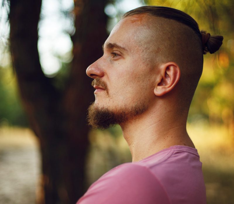 Hipster man met een undercut fade met knot staat in het bos en kruist zijn armen en houdt zijn geiten kin in de lucht.