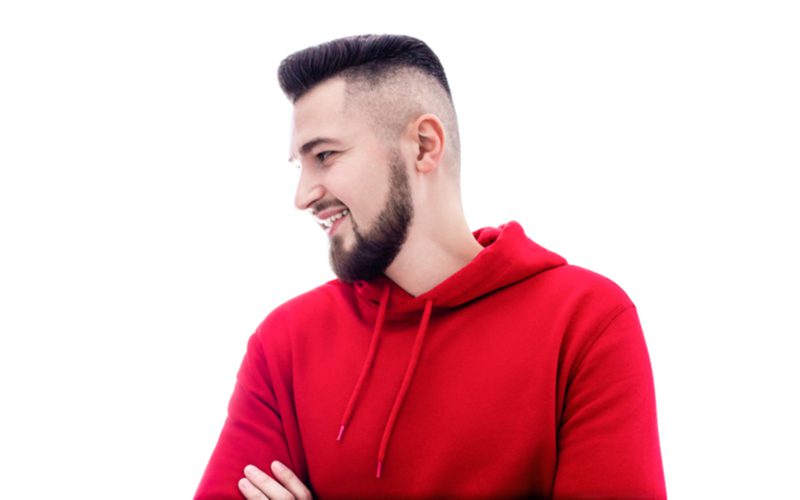 man in een rode hoodie met een hoge undercut fade in een kamer die zijn armen kruist en glimlacht