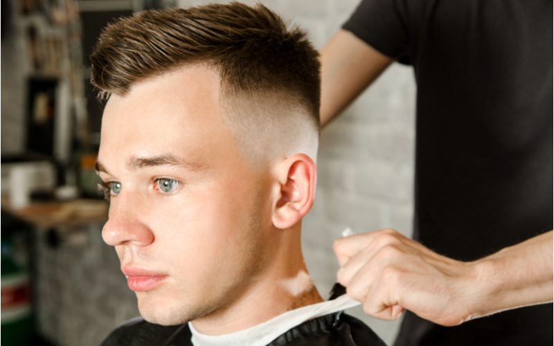 Man met een golvende zijscheiding undercut fade kapsel heeft zijn cape losgeknoopt in een kappersstoel en kijkt voor zich uit zonder te glimlachen