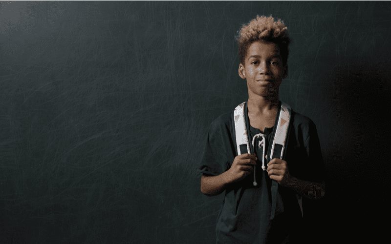 Beeld van een Afro-Amerikaanse jongen die met een rugzak staat terwijl hij de riemen vasthoudt in een donkere kamer