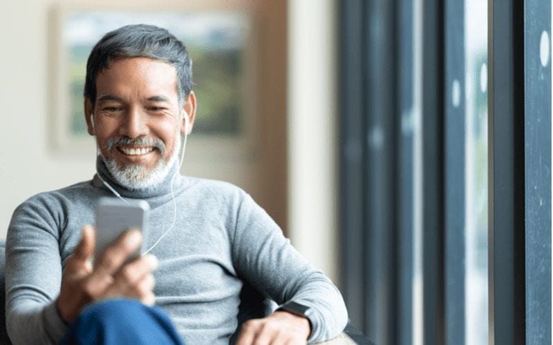 Man met een French Crop kapsel in een coltrui kijkt naar zijn telefoon en lacht in een koptelefoon