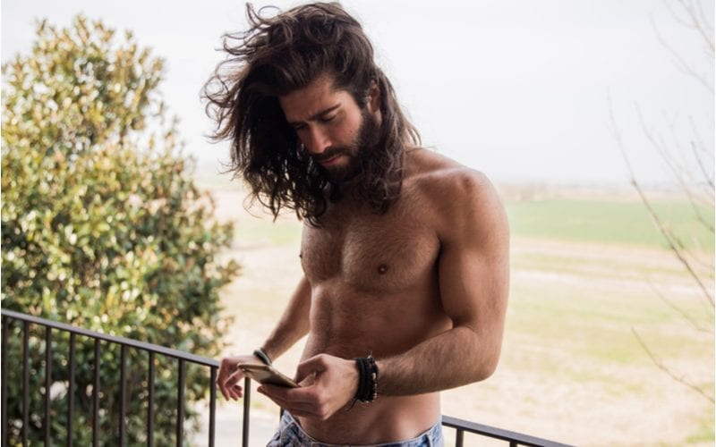 Sexy man zonder tshirt die op een balkon voor een grasveld staat laat zijn lange haar wapperen in de wind