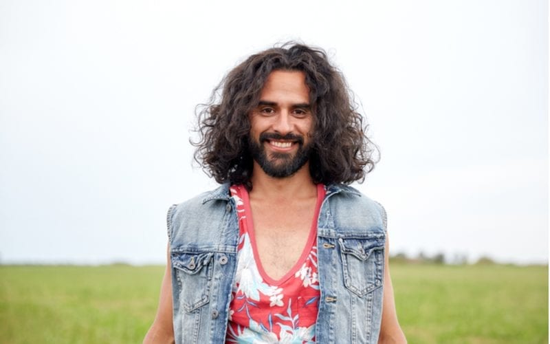 Voor een roundup op lange kapsels voor mannen, draagt een Russell Brand lookalike een bloemrijke tank top en een jeansvest en lacht hij breeduit in een maïsveld.