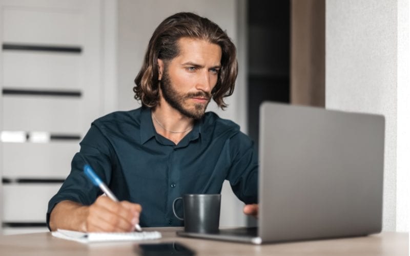 Man met lang haar voor mannen schrijft op een tablet papier terwijl hij naar een laptop kijkt