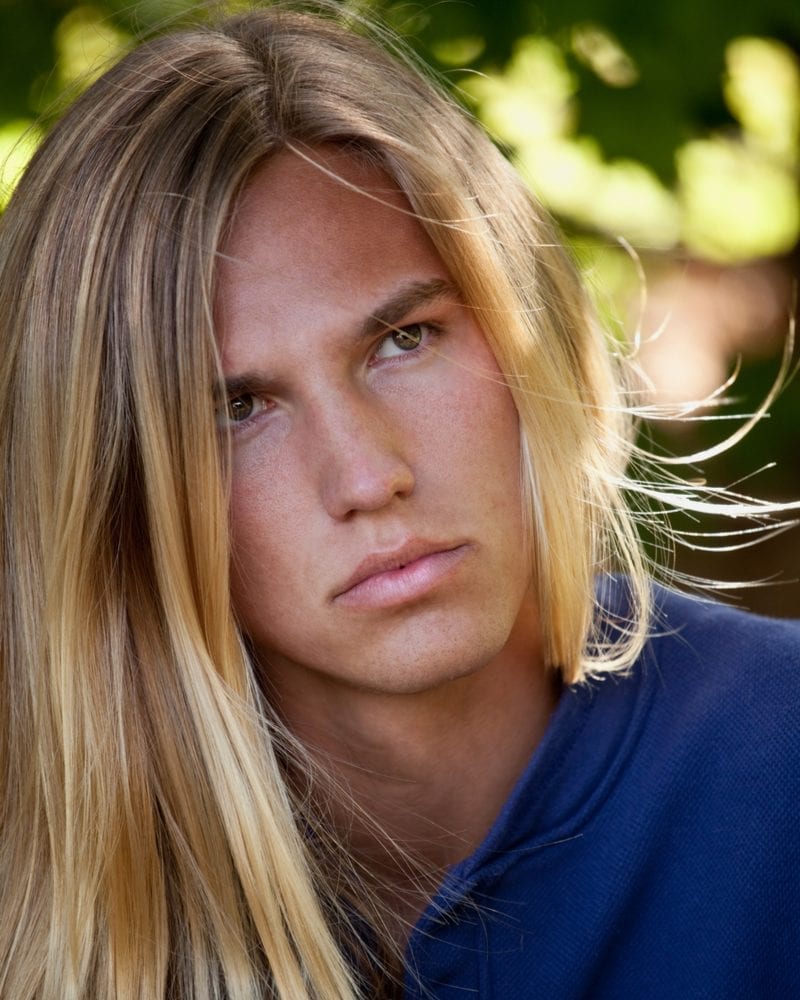 Man met lang haar en een blowout en balayage 90s kapsel die zijn hoofd schuin houdt terwijl hij naar iemand achter de camera luistert.