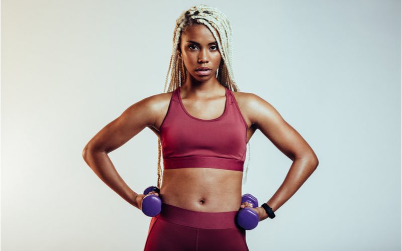 Atletische zwarte vrouw met een scherp kapsel, gebleekte box braids terwijl ze in een sportbeha en yogabroek staat met haar handen op haar heupen.