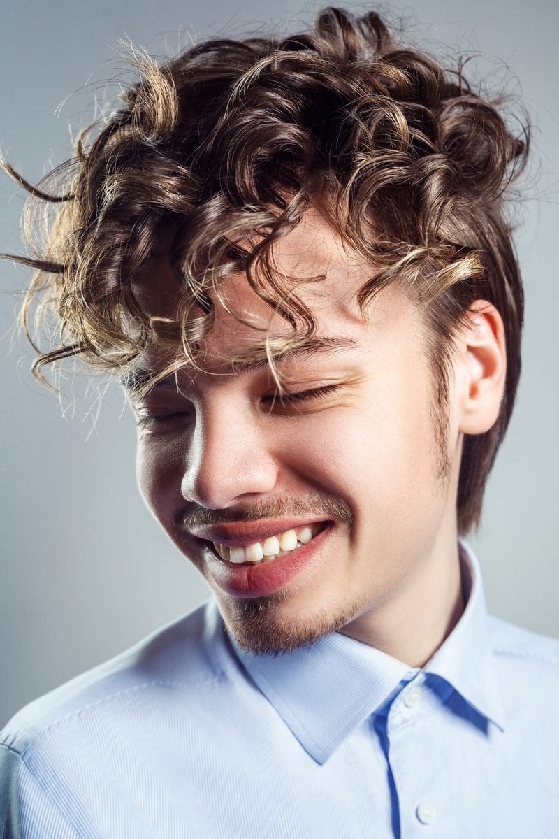 Man met een prins-achtige vetkuif in een blauw overhemd met het bovenste knoopje dicht kijkt naar rechts.