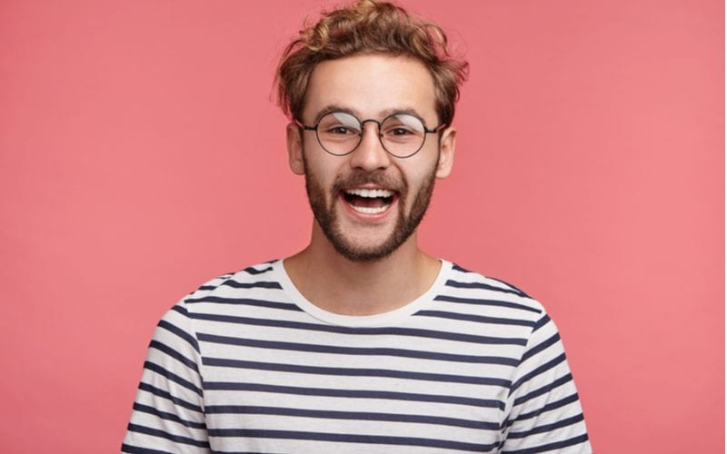 Golvend haar man in een gestreept blauw en wit shirt met ronde hals staat voor een zalmroze muur.