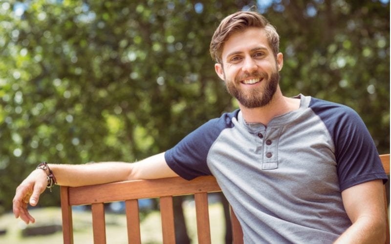 Golvend haar man in een baseball shirt met korte mouwen zit nonchalant op een parkbankje met zijn rechterarm hangend op de rug van het bankje