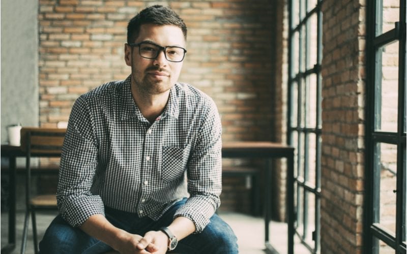 Voor een stuk over de beste kapsels voor mannen met een gevonden gezicht, klemt een man in een gingham shirt zijn handen samen in een stenen kamer