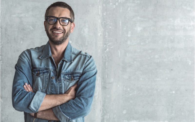 Rond gezicht man met een korte buzz cut glimlacht en draagt een bril terwijl hij zijn armen kruist in een blauw jeans shirt.