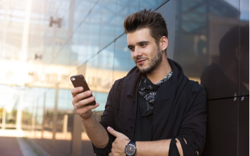 Bebaarde man met een op-en-rug kapsel voor mannen met een rond gezicht kijkt naar zijn telefoon terwijl hij tegen een glazen muur leunt.