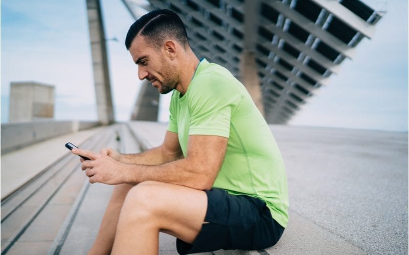 Man gekleed in atletische kleding en een hoge drop fade met geschoren deel kijkt naar zijn telefoon terwijl hij op een betonnen trede zit.