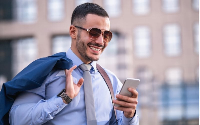 Glimlachende man met een fade buzz cut kijkt naar zijn telefoon en houdt zijn colbert boven zijn rechterschouder