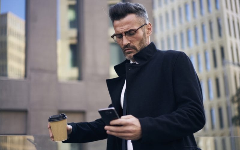 Man in een erwtenjas houdt een koffie in zijn rechterhand en een telefoon in zijn linker terwijl hij buiten een paar hoge gebouwen loopt.