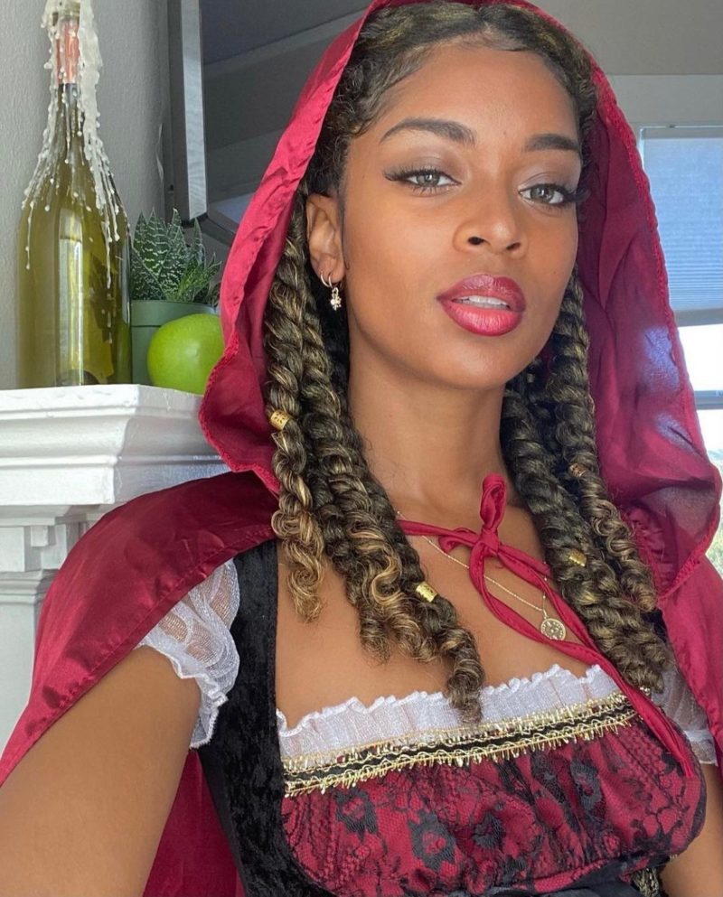 Vrouw met losse natuurlijke kinky twists met een op rood rijdende kap geïnspireerde outfit met de kap omhoog staat voor een schouw.