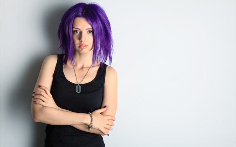 Portret van een meisje met fel paars emo haar dat haar armen kruist in een zwart shirt en tegen een muur leunt