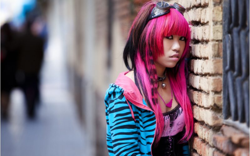 Roze harige Aziatische vrouw met emo haar draagt een blauwe zebra hoodie en een strak spandex topje staande tegen een stenen muur.