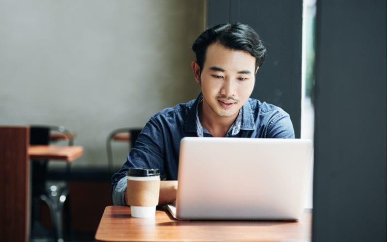 Jonge Aziatische man met een laptop en een koffie op tafel kijkt naar zijn computer in een koffieshop