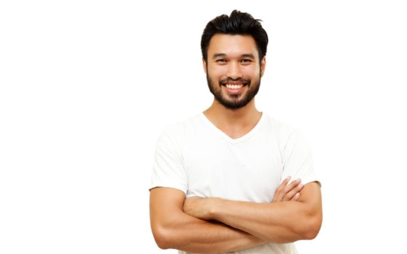 Aziatische man met een Aziatisch mannenkapsel die zijn armen kruist en naar de camera glimlacht in een witte v hals