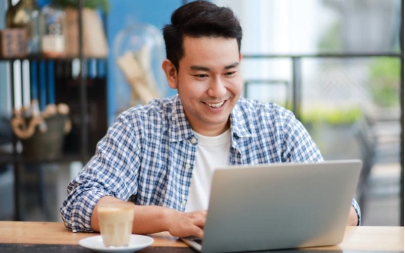 Aziatische man in een losgeknoopt shirt met lange mouwen, lachend en kijkend naar een laptop