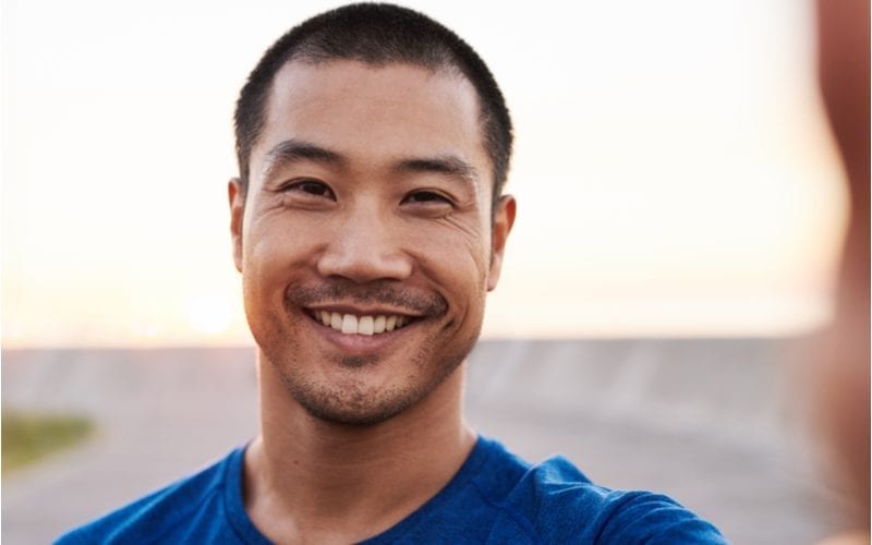 Jonge man glimlacht naar de camera terwijl hij op een strand staat en een blauw T-shirt draagt