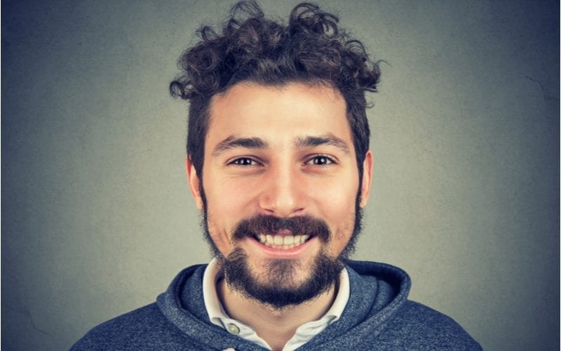 Man met een onverzorgde baard en een gekrulde top kom snit lacht groot naar de camera tegen een grijs verloop achtergrond