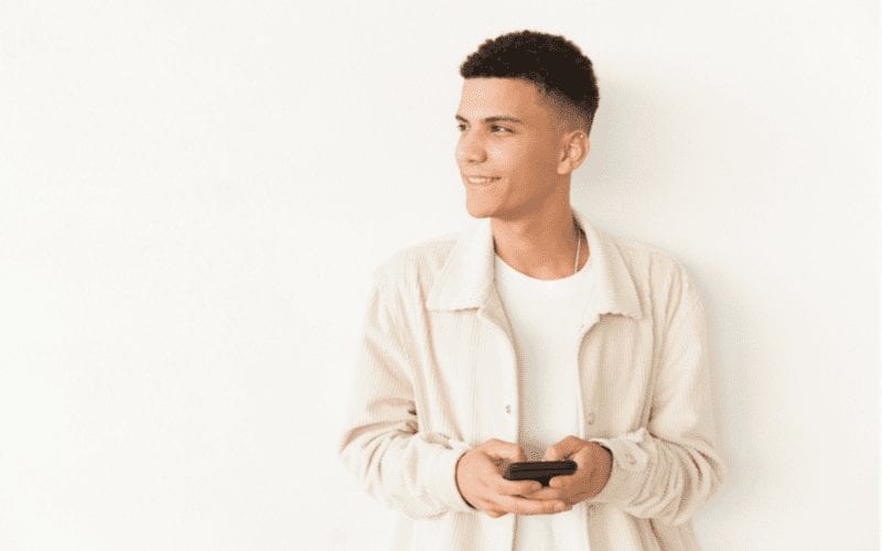 Glimlachende man in een wollen button-up sweater houdt een telefoon vast en denkt na terwijl hij naar rechts kijkt en een komvormige snit draagt.