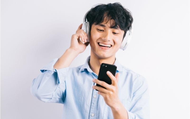 Jonge Aziatische man die glimlacht en naar zijn telefoon kijkt en een koptelefoon vasthoudt die bovenop zijn lange komvormige coupe rust.