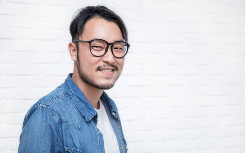Hipster Aziatische man met een gedeelde kom snede staat tegen een blok achtergrond en draagt een open jean jas met wit tshirt
