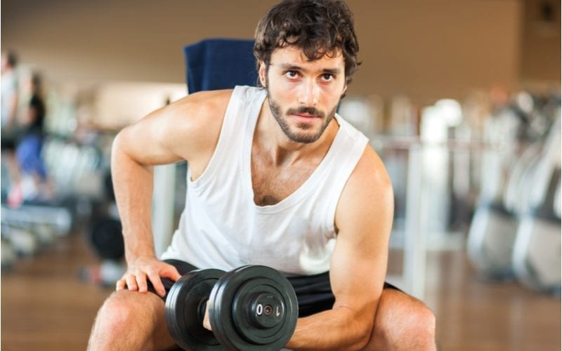 Man met een sportief shag kapsel tilt gewichten op in de sportschool en draagt een tank top.