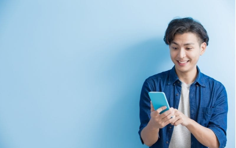 Jonge student houdt zijn telefoon vast en drukt opwinding uit terwijl hij tegen blauwe achtergrond staat