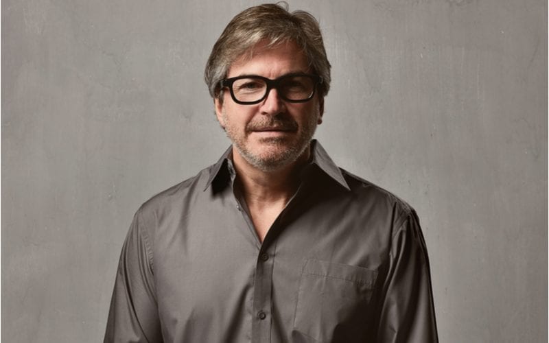 Man in een grijs glimmend overhemd steekt zijn handen in zijn zakken en kijkt door zijn dikgerande bril naar de camera