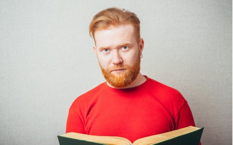 Man met een rood hemd en een roodharig kapsel leest een boek en staat tegen een grijze achtergrond