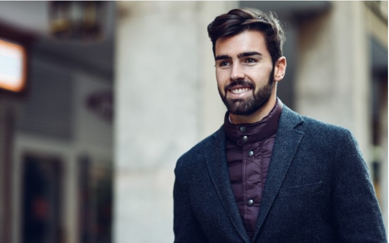 Als afbeelding voor een stuk over een kapsel voor mannen met een groot voorhoofd, draagt een man een Brits uitziend overhemd en wollen jas terwijl hij glimlacht en naar buiten loopt.