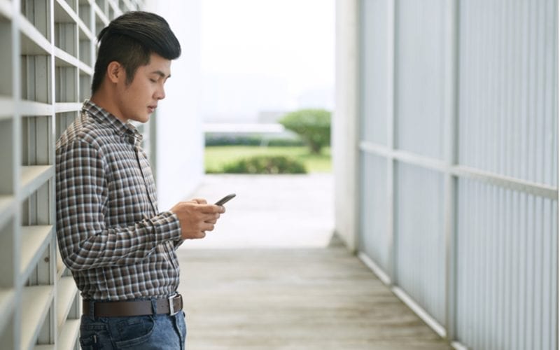Zijprofiel van een jonge Aziatische man staand in een magazijn met zijn hemd ingestopt en kijkend naar een telefoon