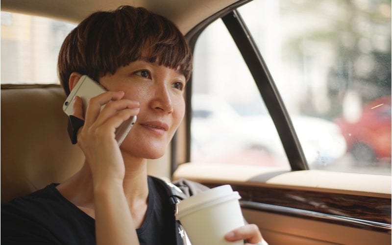 Aziatische vrouw met een kapsel van twee blokken houdt een koffie vast en kijkt voor zich uit in de taxi waarin ze zit