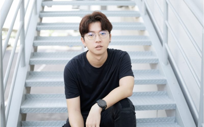 Aziatische man met bril en two block kapsel zit op de trappen van een gebouw en kijkt naar de camera