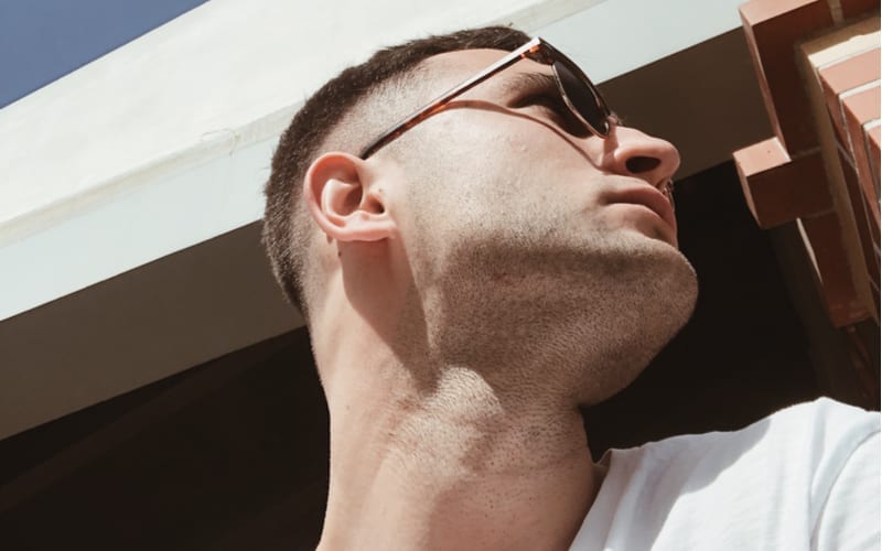 Selfie van een stijlvolle en aantrekkelijke man in profiel die een wit t-shirt en een zonnebril draagt. Knappe jongeman met zonnebril en stoppelbaard die opzij kijkt onder zonlicht. Jongen met trendy fade undercut kapsel
